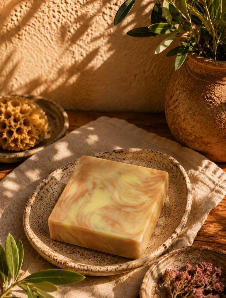 Kaltgerührte Manufakturseife Village Bloom von PACATS COSMETIC MANUFACTURE in einer Keramikschale. Das stimmungsvolle Foto zeigt die Naturseife mit Mustern aus Tonerde im warmen, mediterranen Tageslicht, kunstvoll dekoriert mit einem echten Naturschwamm, getrockneten Blüten und einem Terrakottakrug mit Olivenblättern auf einem Holztisch.