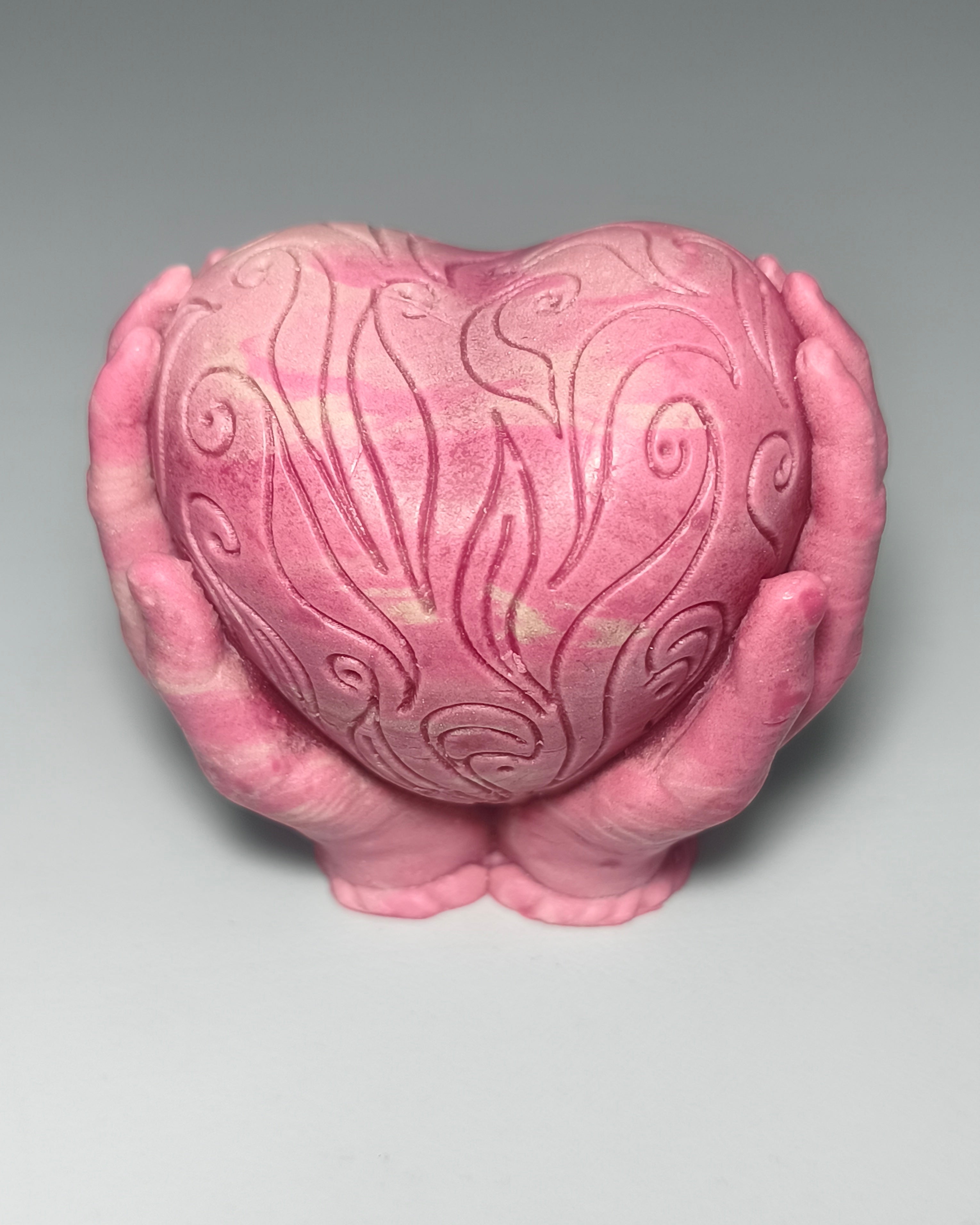 Handgemachte rosa Seife in Form eines verzierten Herzens, das von zwei Händen gehalten wird, mit feinen Schnörkel-Schnitzereien und leichter Marmorierung, von PACATS.