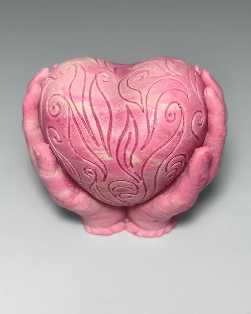 Handgemachte rosa Seife in Form eines verzierten Herzens, das von zwei Händen gehalten wird, mit feinen Schnörkel-Schnitzereien und leichter Marmorierung, von PACATS.