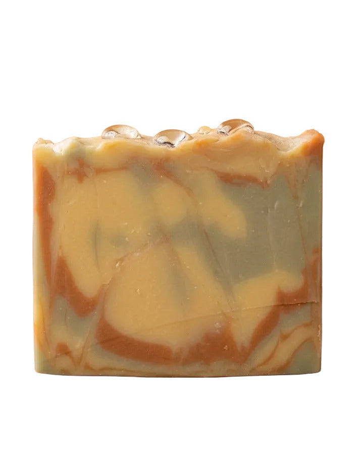 Ein eckiges Stück handgemachte Naturseife Oil & Clay vor einem einfarbigen, warmen terracotta-braunen Hintergrund. Das Seifenstück zeigt ein dreifarbiges, marmoriertes Swirl-Muster aus gelblichen, blau-grauen und rötlich-braunen Tonerden. Auf der welligen, rustikalen Oberkante des Seifenstücks liegen drei klare, glänzende Wassertropfen.