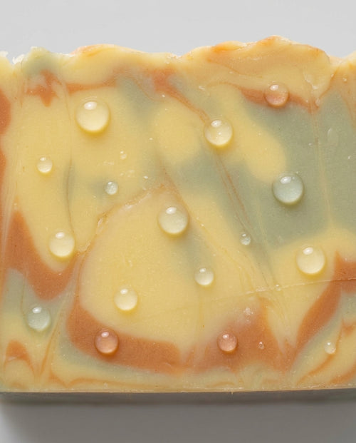 Eine Makro-Nahaufnahme der Oberfläche eines rechteckigen Seifenstücks Oil & Clay vor einem hellgrauen Hintergrund. Auf dem dreifarbigen, kunstvoll marmorierten Swirl-Muster aus gelblichen, blau-grauen und rötlich-braunen Tönen liegen zahlreiche, perfekt runde und glänzende Wassertropfen verstreut. Die Textur der glatten Seifenoberfläche und die Lichtreflexionen in den Tropfen sind deutlich sichtbar.