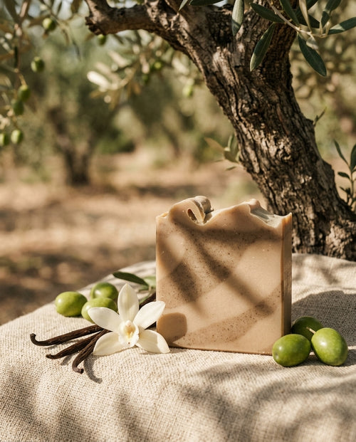 Ein Stück rechteckige Naturseife Olive-Vanilla steht auf einer hellen Jutedecke im Schatten eines Olivenbaums. Neben der Seife liegen frische grüne Oliven, getrocknete Vanilleschoten und eine weiße Vanilleblüte. Das Sonnenlicht wirft sanfte Blätterschatten auf das Seifenstück mit erdigen, braun-beigen Mustern.