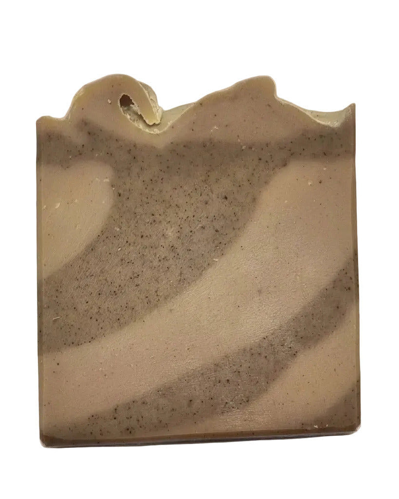 Handgefertigtes, rechteckiges Stück Naturseife mit einer unregelmäßigen, welligen Oberkante. Das Seifenstück zeigt ein sanftes, wellenförmiges Muster aus abwechselnd hellbeigen und dunkelbraun gesprenkelten Erdtönen vor einem einfarbigen, hellbraunen Hintergrund.